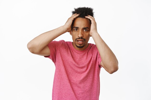 Frustrierter schockierter Afroamerikaner, der die Hände auf dem Kopf hält, die Stirn runzelt und besorgt in die Kamera starrt, verängstigt oder alarmiert über etwas, das in rosa T-Shirt vor weißem Hintergrund steht