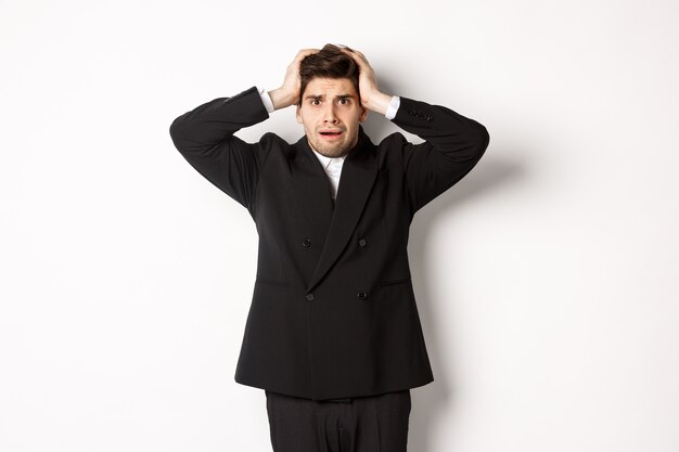 Frustrierter und besorgter Geschäftsmann im schwarzen Anzug, der in Panik verfällt, als er Ärger betrachtet, die Hände alarmiert am Kopf hält und vor weißem Hintergrund steht
