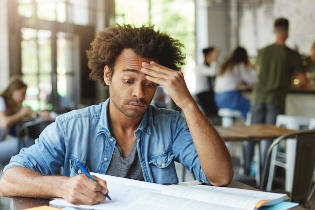 Frustrierter verwirrter junger Student mit Afro-Frisur, die sich die Stirn reibt und sich bemüht, komplizierte mathematische Probleme zu verstehen, während er Hausaufgaben im Café macht und mit dem Stift Notizen macht