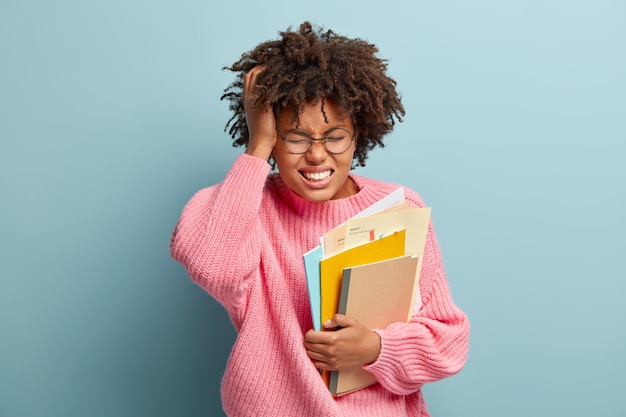 Frustriertes schulmädchen hat schreckliche kopfschmerzen, runzelt die stirn vor schmerzen, lernt gut, führt tagebuch mit papieren, trägt eine runde optische brille und einen rosa pullover, steht über einer blauen wand und ist traurig, den test vergessen zu haben