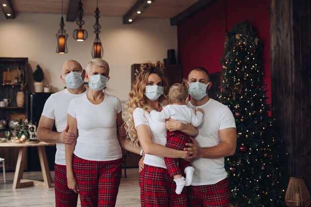 Fünf Familienmitglieder mit einem Baby in T-Shirts und Hosen in schützenden Gesichtsmasken zu Weihnachten zu Hause.