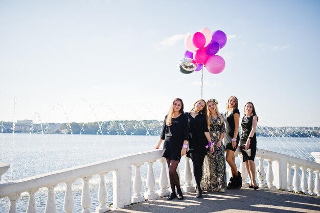 Fünf Mädchen tragen beim Junggesellinnenabschied gegen den See schwarze Kleidung, die mit Luftballons spazieren geht