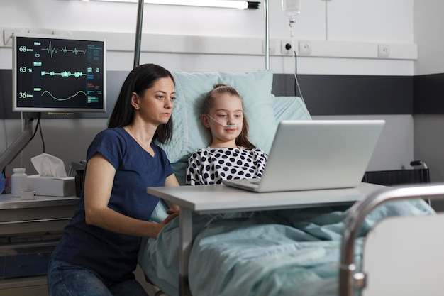 Fürsorgliche Mutter, die neben der kranken Tochter in Behandlung sitzt, während sie gemeinsam Cartoons auf dem Laptop in der Kinderstation des Krankenhauses anschaut. Krankes Kind und liebevolle Eltern genießen die gemeinsame Zeit in der Klinik.