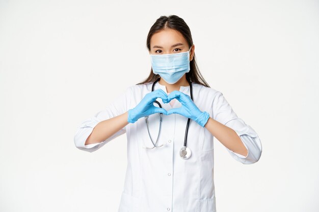 Fürsorglicher asiatischer Arzt, Ärztin in medizinischer Maske und Gummihandschuhen zeigt ihre Pflege für Patienten mit Herzzeichen, weißem Hintergrund