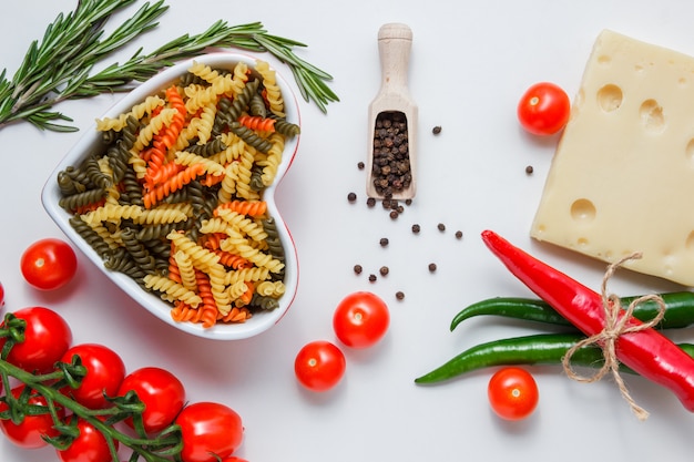 Fusilli Nudeln in einer Schüssel mit Paprika, Tomaten, Käse, Pflanze, Pfefferkörnern flach auf einem weißen Tisch liegen