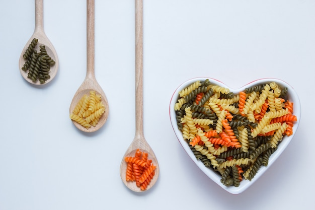 Fusilli Nudeln in Schüssel und Holzlöffel auf weißem Tisch, Draufsicht.