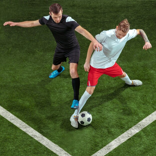 Fußballspieler, der für Ball über Hintergrund des grünen Grases angreift. Professionelle männliche Fußballspieler in Bewegung im Stadion. Fit springende Männer in Aktion, Sprung, Bewegung beim Spiel.