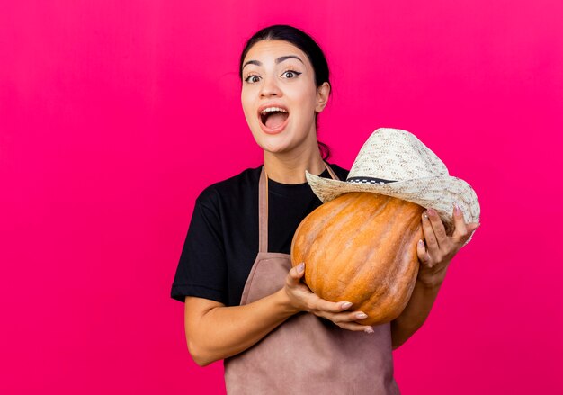 Gärtnerin der jungen schönen Frau in der Schürze, die Pumpking mit dem lächelnden Hut hält, der Spaß hat