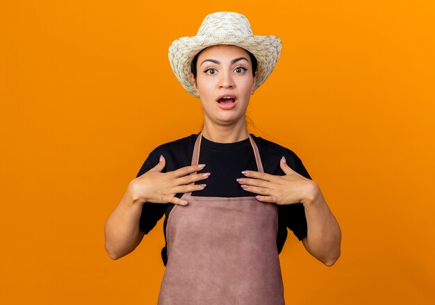 Gärtnerin der jungen schönen Frau in der Schürze und im Hut, die auf sich selbst zeigen, die über orange Wand stehen überrascht sind