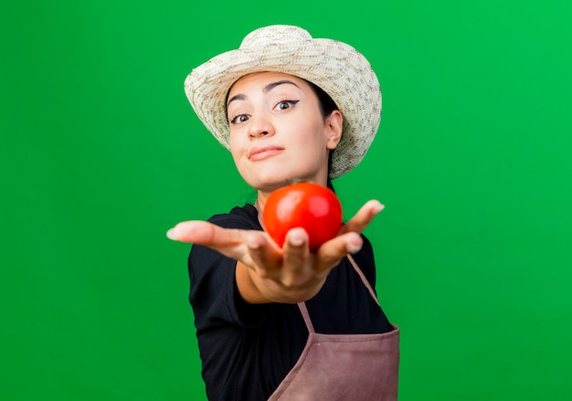 Gärtnerin der jungen schönen Frau in der Schürze und im Hut, die das lächelnde selbstbewusste Stehen der Tomate über der grünen Wand zeigen