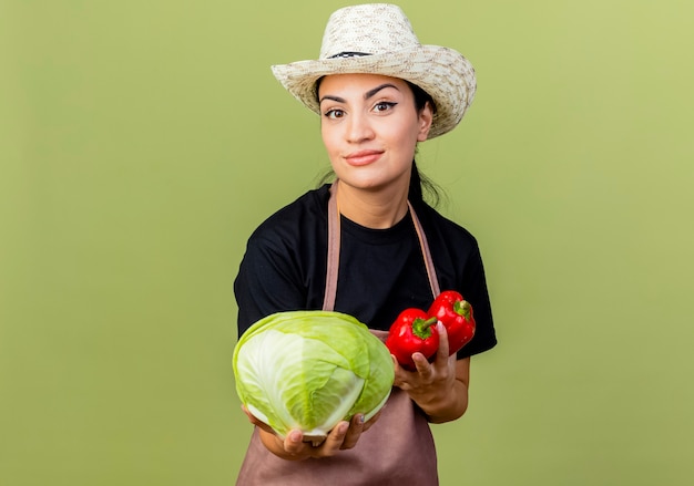 Gärtnerin der jungen schönen Frau in der Schürze und im Hut, die Kohl und rote Paprika halten, die über hellgrüner Wand lächelnd stehen