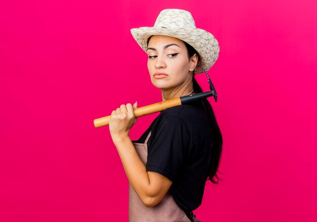 Gärtnerin der jungen schönen Frau in der Schürze und im Hut, die Mini-Rechen halten, der beiseite mit ernstem Gesicht steht über rosa Wand steht