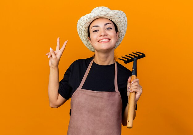 Gärtnerin der jungen schönen Frau in der Schürze und im Hut, die Mini-Rechen halten, der vorne lächelnd zeigt, das V-Zeichen zeigt, das über orange Wand steht