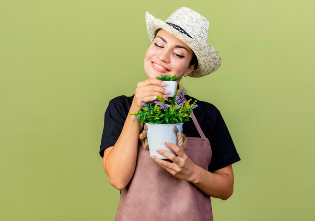 Gärtnerin der jungen schönen Frau in der Schürze und im Hut, die Topfpflanzen halten, die es lächelnd mit glücklichem Gesicht betrachten, das über hellgrüner Wand steht