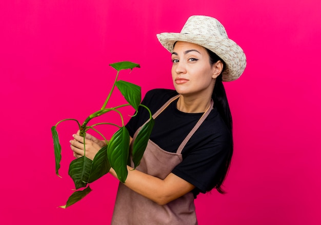 Gärtnerin der jungen schönen Frau in der Schürze und in der Hut, die Pflanze hält, die vorne mit skeptischem Ausdruck steht, der über rosa Wand steht