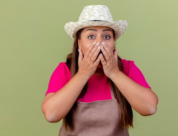 Gärtnerin mittleren Alters in der Schürze und im Hut, die Kamera betrachten, die erstaunt und überrascht ist, Mund mit den Händen zu bedecken, die über hellem Hintergrund stehen