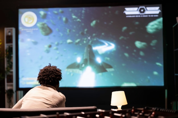 Kostenloses Foto gamer vor einem gigantischen smart-tv beobachtet content-schöpfer, der während des livestreams ein raumschiff in der galaxie pilotiert