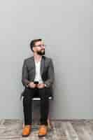 Kostenloses Foto ganzaufnahme des entspannten mannes im zufälligen sitzen auf stuhl im büro, das beiseite mit smartphone in den händen, lokalisiert über grau schaut