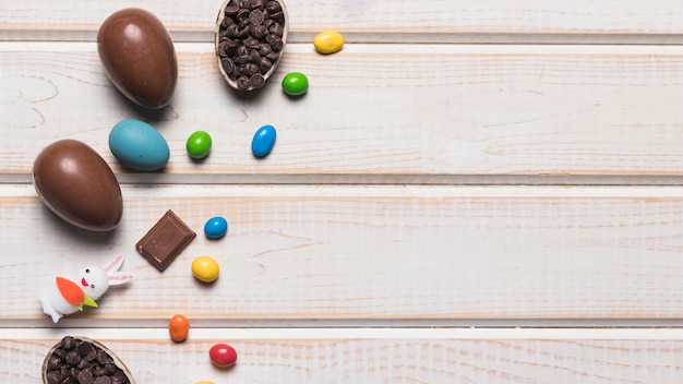 Ganze Ostereier aus Schokolade bunte Edelstein-Bonbons; Choco-Chips und Kaninchen auf Schreibtisch aus Holz