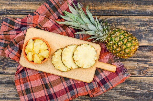 Ganze und geschnittene Ananas in einem Schneidebrett und einer Tonschale auf einer Holz-Grunge-Oberfläche und einem Picknicktuch. flach liegen.