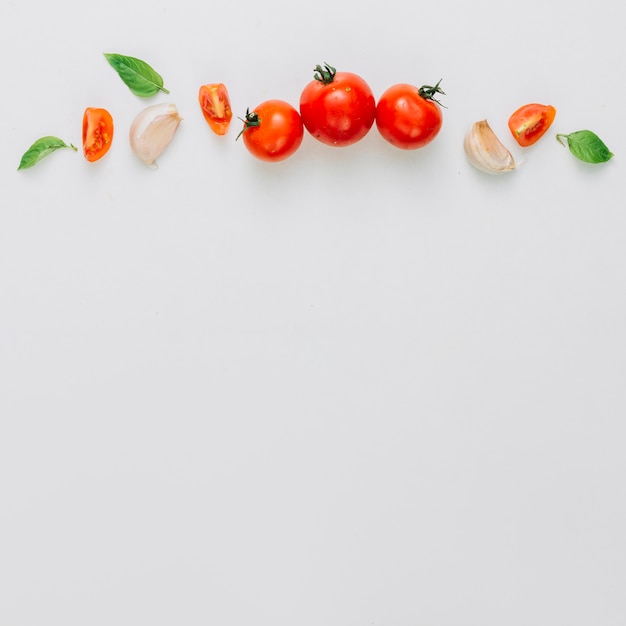 Ganze und in Scheiben geschnittene Kirschtomaten; Knoblauchzehe und Basilikum über dem weißen Hintergrund