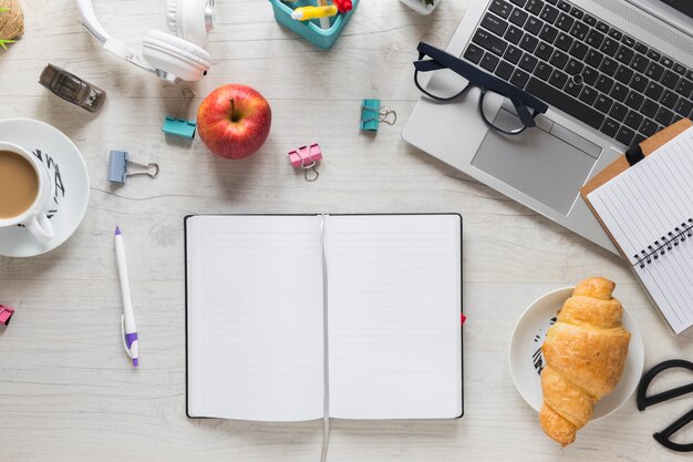 Ganzer roter Apfel; Croissant und Teetasse mit Bürobedarf und Laptop auf Holztisch