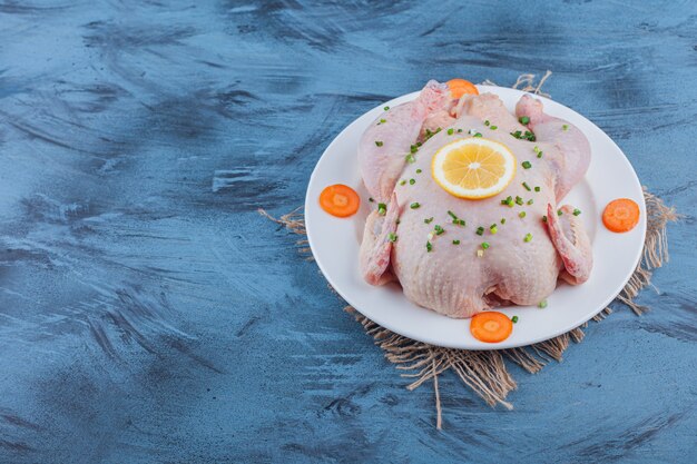 Ganzes Huhn, Zitrone und geschnittene Karotten auf einem Teller auf einer Leinwand, auf dem blauen Tisch.