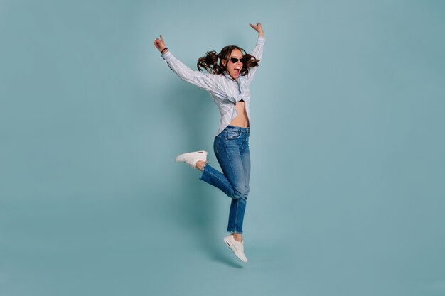 Ganzes Porträt des lustigen reizenden weiblichen Modells, das springt und Spaß über isolierter Wand hat. Glückliches Mädchen tanzt und lächelt auf blauem isoliertem Hintergrund.