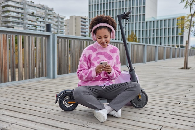 Ganzkörperaufnahme einer hübschen fröhlichen Teenagerin sitzt mit gekreuzten Beinen und nutzt das Smartphone, um online zu chatten und Musik über Kopfhörer zu hören, nachdem sie mit dem Elektroroller durch die Stadt gefahren ist