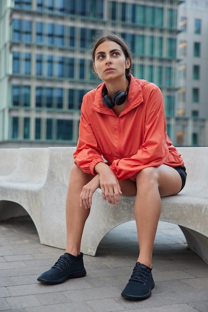 Ganzkörperaufnahme einer nachdenklichen europäischen Sportlerin macht Pause, nachdem das in Activewear gekleidete Cardio-Training auf Stein gegen die Stadt sitzt