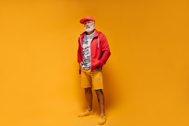 Ganzkörperaufnahme eines Mannes in orangefarbenen Shorts und roter Jacke Stylischer Typ mit Bart in stylischem Hoodie mit weißem Leopardenmuster und Mütze posiert