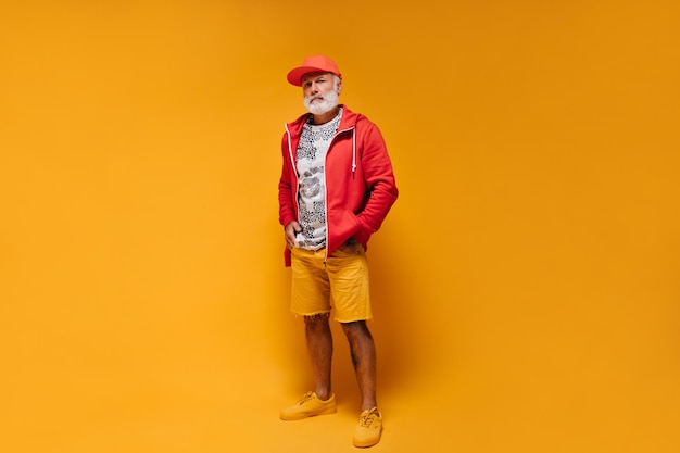 Ganzkörperaufnahme eines Mannes in orangefarbenen Shorts und roter Jacke Stylischer Typ mit Bart in stylischem Hoodie mit weißem Leopardenmuster und Mütze posiert