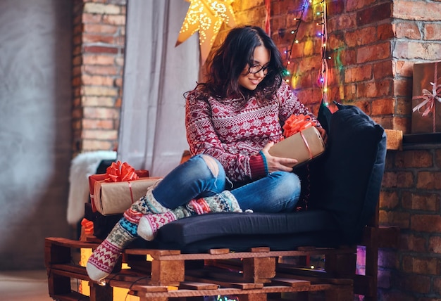 Ganzkörperbild einer brünetten Frau mit Brille in Jeans und rotem Pullover, die auf einem Holzsofa in einem Raum mit Weihnachtsdekoration posiert.