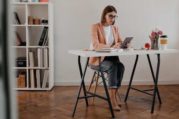 Ganzkörperfoto einer Geschäftsfrau in rosa Jacke und Jeans, die in ihrem Büro mit dem iPad arbeitet