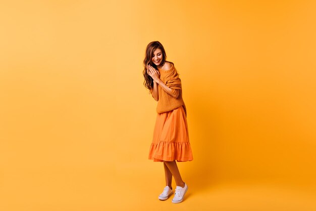 Ganzkörperfoto einer süßen jungen Frau in weißen Gummischuhen isoliert auf gelbem Hintergrund Studioporträt eines wunderschönen dunkelhaarigen Mädchens in orangefarbenem Outfit