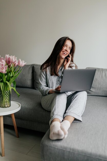 Ganzkörperporträt einer aufgeregten, schönen Frau mit dunklem Haar, die einen Schlafanzug trägt, spricht am Telefon und arbeitet zu Hause mit dem Laptop Frau zu Hause