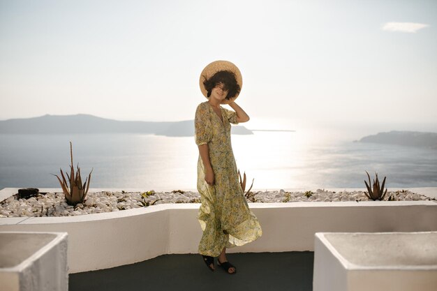 Ganzkörperporträt einer glücklichen brünetten Dame im Midikleid und Bootsfahrer, die auf der Terrasse mit Meerblick posieren Attraktive Frau mit Hut und Blumenoutfit lächelt auf Meereshintergrund