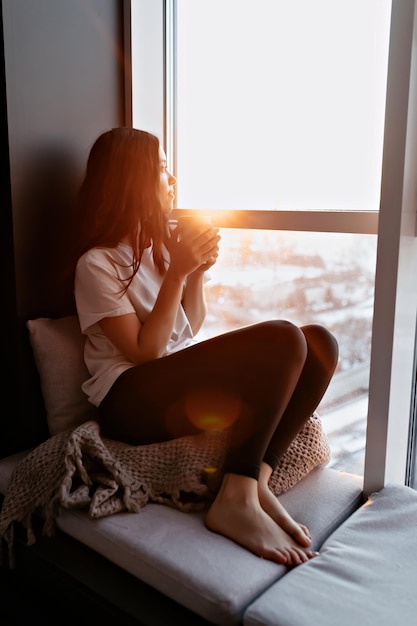 Ganzkörperporträt einer jungen hübschen Dame mit weißem T-Shirt und Hosen sitzt im Sonnenlicht neben dem Fenster mit Kaffee und blickt auf die Stadt