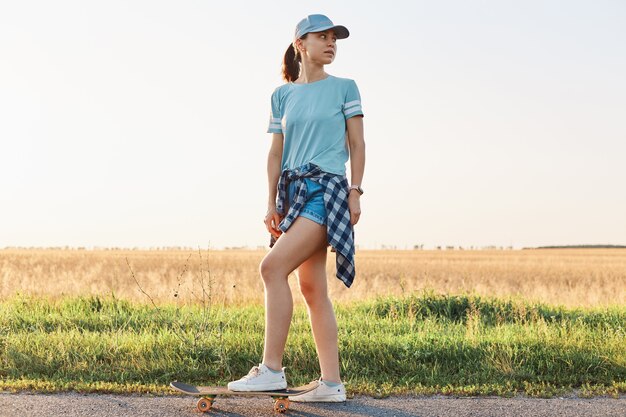 Ganzkörperporträt einer schlanken, sportlichen Frau mit T-Shirt und Schirmmütze, die mit dem Bein auf dem Skateboard steht und wegschaut, ihre Freizeit aktiv verbringt.