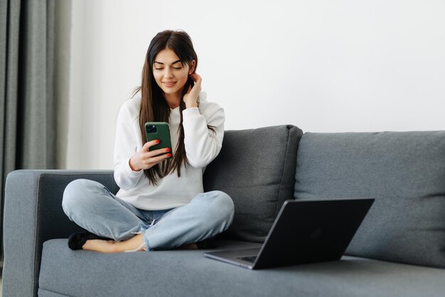 Ganzkörperporträt einer sorglosen Frau, die ihr Handy hält und mit Lächeln und Freude auf der heimischen Couch kommuniziert