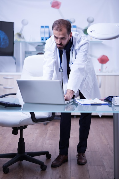 Ganzkörperporträt eines gutaussehenden Arztes, der an seinem Laptop im Krankenhausschrank arbeitet. Arzt in einem Labor.