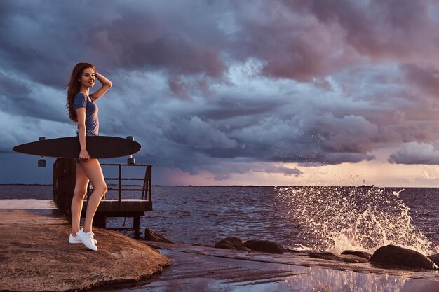 Ganzkörperporträt eines sinnlichen Mädchens hält ein Skateboard, während es am Strand steht und während des Sonnenuntergangs erstaunliches dunkles bewölktes Wetter genießt.