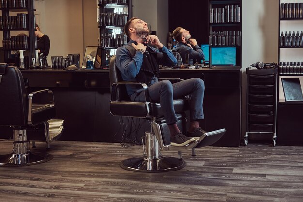 Ganzkörperporträt eines stilvollen Mannes, der sich selbst rasiert, während er auf einem Friseurstuhl in einem Friseursalon sitzt.