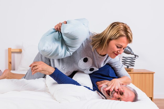 Gealterte Frau auf dem Mann, der Spaß mit Kissen hat und auf Bett liegt