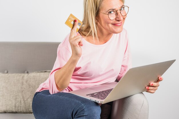 Gealterte lächelnde Frau in der rosafarbenen Bluse, die Plastikkarte zeigt und Laptop auf Sofa verwendet