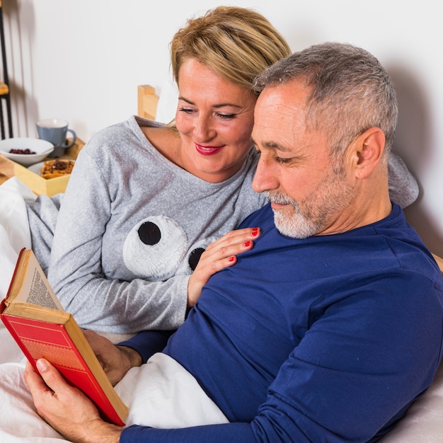 Gealterte lächelnde Frau nahe Mann mit Buch in der Daunendecke auf Bett