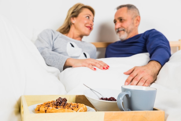 Gealterter lächelnder Mann und Frau in der Daunendecke nahe Frühstück auf Tellersegment auf Bett