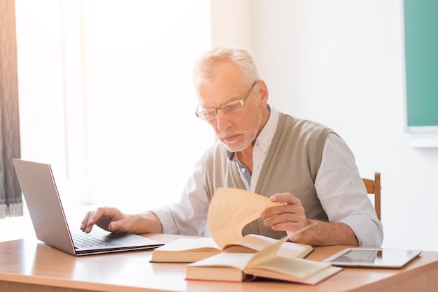 Gealterter Professormann, der mit Laptop beim Lesebuch im Klassenzimmer arbeitet