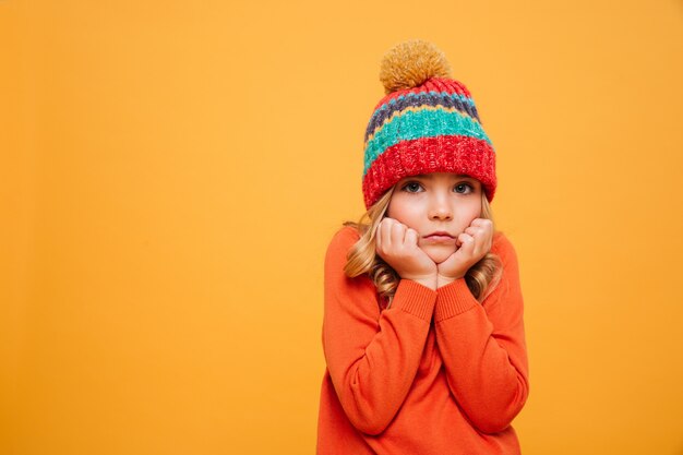 Gebohrtes junges Mädchen in der Strickjacke und im Hut stützt sich auf ihre Arme und betrachtet die Kamera über Orange