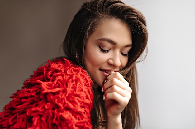 Gebräunte attraktive Dame im roten ungewöhnlichen Pullover lächelt mit geschlossenen Augen auf weißem Hintergrund.